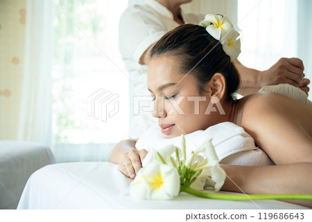Young woman with eyes closed, receiving herbal compress massage at spa bed, resting on towel with fresh flowers nearby 119686643