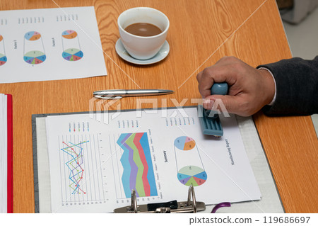 Close-up of businessman stamping financial report with charts and graphs on desk, next to cup of coffee and pen, in office Close-up of businessman stamping financial report with charts and graphs on desk, next to cup of coffee and pen, in office 119686697