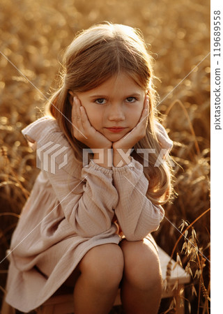 A young blonde girl in a beige dress sits in a field with tall grass. A young blonde girl in a beige dress sits in a field with tall grass. 119689558
