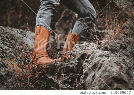 Human feet in red cowboy boots on a rock among beautiful atmospheric autumn herbs outdoor, close-up, selective focus. 119689697