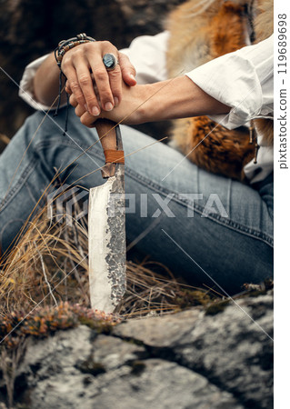 Rude handmade knife in female hands, faceless woman in fox fur vest sits on a rock outdoor, vertical format, selective focus. Rude handmade knife in female hands, faceless woman in fox fur vest sits on a rock outdoor, vertical format, selective focus. 119689698