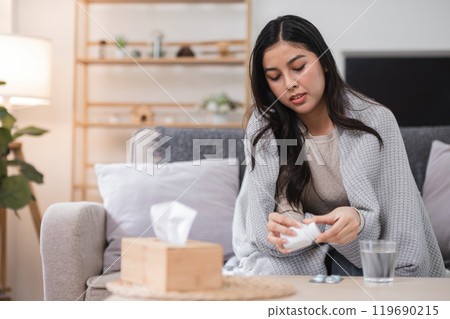 Young Woman Feeling Unwell on Sofa at Home, Wrapped in Blanket and Taking Medication 119690215