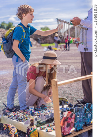 Mother and son tourists buying Mexican souvenirs from a street vendor at the foot of the Teotihuacan pyramid. Cultural experience, travel, and local craftsmanship concept Mother and son tourists buying Mexican souvenirs from a street vendor at the foot of the Teotihuacan pyramid. Cultural experience, travel, and local craftsmanship concept 119691819