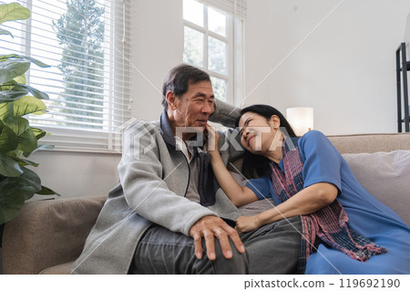 Elderly Couple Relaxing in Cozy Living Room, Enjoying Quality Time Together, Smiling and Embracing, Surrounded by Modern Home Decor and Natural Light from Large Windows 119692190