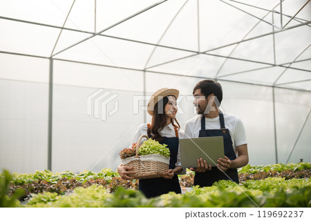 Happy Couple Engaged in Modern Agriculture Inside a Greenhouse, Holding Fresh Produce and a Tablet 119692237