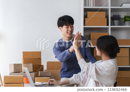 Happy Couple Running a Small Online Business from Home, Surrounded by Cardboard Boxes and Working on a Laptop, Celebrating Success with a High Five in a Bright Modern Workspace Happy Couple Running a Small Online Business from Home, Surrounded by Cardboard Boxes and Working on a Laptop, Celebrating Success with a High Five in a Bright Modern Workspace 119692466