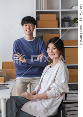 Young Couple Running a Small Online Business from Home, Surrounded by Shipping Boxes and Office Supplies, Smiling and Confident in Their Modern Workspace 119692468