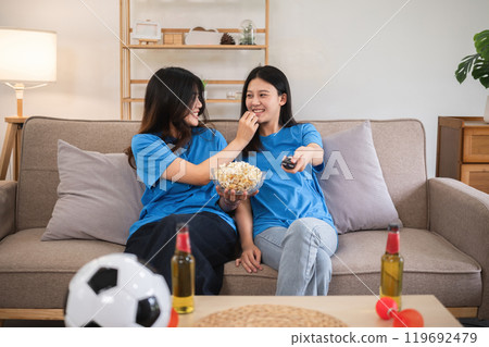 Lesbian Couple Cheering for Football Together on Couch, Enjoying Snacks and Drinks, Celebrating Sports Enthusiasm and Love, Cozy Home Setting, LGBTQ Representation, Happy Moments, Casual Clothing 119692479
