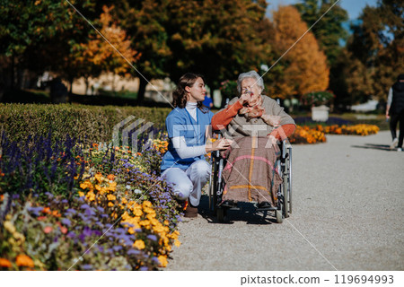 Disabled elderly woman in wheelchair on walk in the park during warm autumn day. Young nurse showing senior woman a flower. 119694993