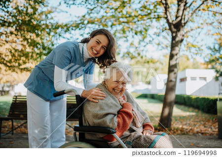 Disabled elderly woman in wheelchair on walk in the park during warm autumn day. Young nurse pushing wheelchair, talking with senior woman. Disabled elderly woman in wheelchair on walk in the park during warm autumn day. Young nurse pushing wheelchair, talking with senior woman. 119694998
