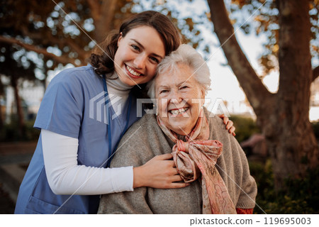 Portrait of nurse and elderly woman on walk in the park during warm autumn day. Young caregiver spending time with senior patient. 119695003