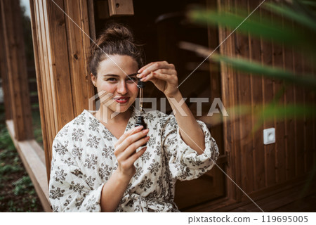 Beautiful woman doing her skin care routine outdoors, wearing bathrobe. Woman using facial skin serum with dropper. Beautiful woman doing her skin care routine outdoors, wearing bathrobe. Woman using facial skin serum with dropper. 119695005