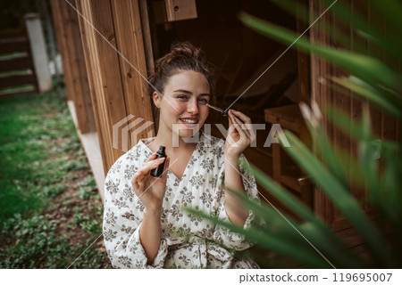 Beautiful woman doing her skin care routine outdoors, wearing bathrobe. Woman using facial skin serum with dropper. Beautiful woman doing her skin care routine outdoors, wearing bathrobe. Woman using facial skin serum with dropper. 119695007