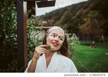 Beautiful woman massaging her face with green jade gua sha tool. Outdoor skin care routine for young woman. Beautiful woman massaging her face with green jade gua sha tool. Outdoor skin care routine for young woman. 119695079