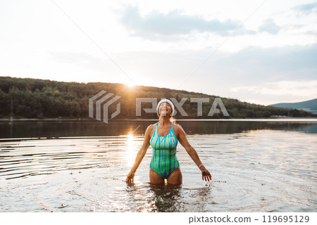 Cold water swimming for elderly women. Portrait of senior sporty women standing in lake during cold evening. Cold water swimming for elderly women. Portrait of senior sporty women standing in lake during cold evening. 119695129