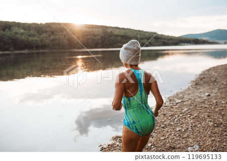 Cold water swimming for elderly woman. Rear view of senior sporty woman wade into lake during cold morning, looking at sunrise. Cold water swimming for elderly woman. Rear view of senior sporty woman wade into lake during cold morning, looking at sunrise. 119695133