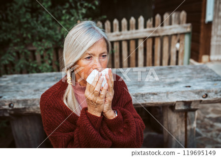Senior women feeling sick, blowing her runny, stuffy nose into tissue and sneezing. 119695149