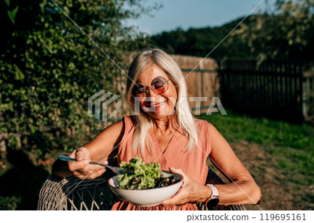 Senior woman is sitting in the garden, eating salad from her own harvest. Older woman is enjoying peaceful lunch alone. Senior woman is sitting in the garden, eating salad from her own harvest. Older woman is enjoying peaceful lunch alone. 119695161