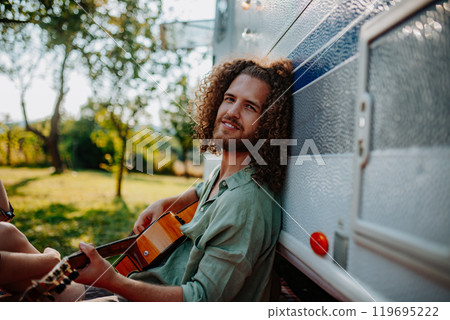 Young man is on a camping trip in nature, sitting in front caravan and playing guitar alone. 119695222