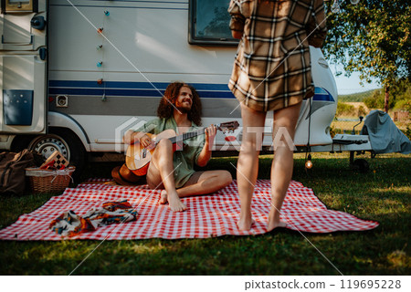 Couple sitting by caravan, enjoying peaceful moment. Man playing guitar and singing song to his girlfriend. Camping trip for young people. Couple sitting by caravan, enjoying peaceful moment. Man playing guitar and singing song to his girlfriend. Camping trip for young people. 119695228