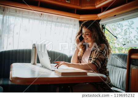 Young beautiful woman is on a camping trip in nature, sitting in her caravan making phone call. Caravan as homeoffice, working remotely. Young beautiful woman is on a camping trip in nature, sitting in her caravan making phone call. Caravan as homeoffice, working remotely. 119695232