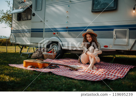 Young beautiful woman is on a camping trip in nature, sitting in front caravan and reading book. 119695239
