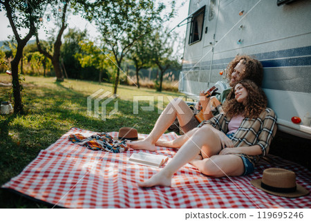 Couple sitting by caravan, enjoying peaceful moment. Man playing guitar and singing song to his girlfriend. Camping trip for young people. Couple sitting by caravan, enjoying peaceful moment. Man playing guitar and singing song to his girlfriend. Camping trip for young people. 119695246