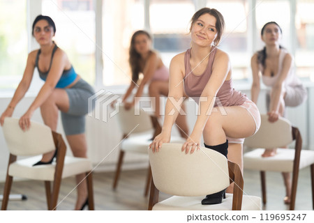 Leaning on chair seat with hand foot, young woman performs lap dance during group workout. 119695247