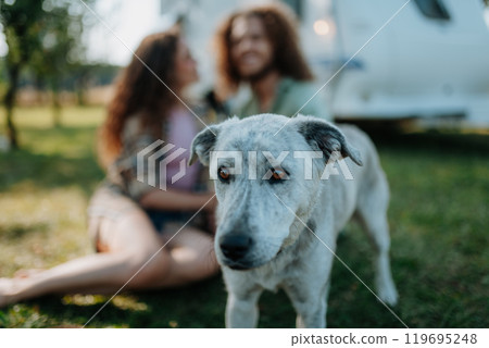 Young couple and dog are on a camping trip in nature, sitting in front of caravan. Caravan traveling for young people. Young couple and dog are on a camping trip in nature, sitting in front of caravan. Caravan traveling for young people. 119695248