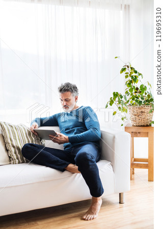 Portrait of attractive middle aged man at home, watching tv show on tablet, sitting on sofa in living room. 119695310