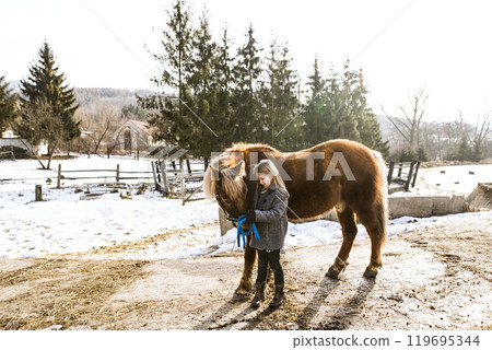 Young girl spending cold winter weekend on a farm, taking care of animals and petting brown fluffy pony. 119695344