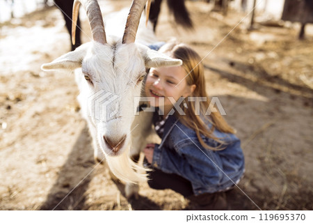 Young girl spending cold winter weekend on a farm, taking care of animals and petting white goat. 119695370
