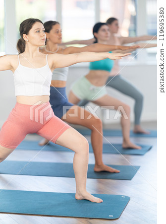 Young girl teaches yoga lesson, shows students technique of performing warrior pose asana Young girl teaches yoga lesson, shows students technique of performing warrior pose asana 119695380