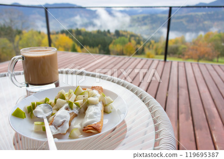 Pancakes with chopped fruits and coffee cup on amazing autumn landscape background. Breakfast in mountains. Breakfast on wooden terrace in mountain resort. Tourism in Ukraine.  119695387
