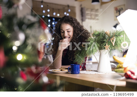 Christmas hygge at home. Beautiful young woman eating christmas cookies and sweets next to the Christmas tree. 119695499