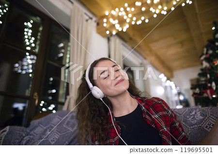 Student having hygge Christmas at home. Woman is sitting on sofa by Christmas tree, listening music with headphones. Student having hygge Christmas at home. Woman is sitting on sofa by Christmas tree, listening music with headphones. 119695501