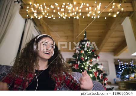 Student having hygge Christmas at home. Woman is sitting on sofa by Christmas tree, listening music with headphones. 119695504