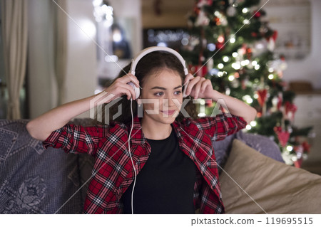 Student having hygge Christmas at home. Woman is sitting on sofa by Christmas tree, listening music with headphones. Student having hygge Christmas at home. Woman is sitting on sofa by Christmas tree, listening music with headphones. 119695515