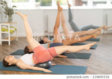 Group of women lying on their backs doing yoga with exercise balls under their backs in studio 119695516