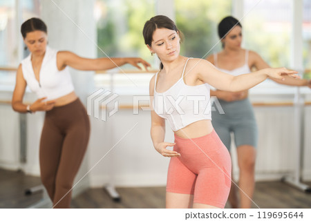 Cheerful active young girl training various ballet poses during group classes with ballet dancers, rehearsal before performance in choreography studio 119695644