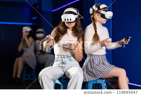 Girls wearing virtual reality glasses play in pairs, against each other, manipulating objects or shooting at targets using joysticks in their hands 119695647