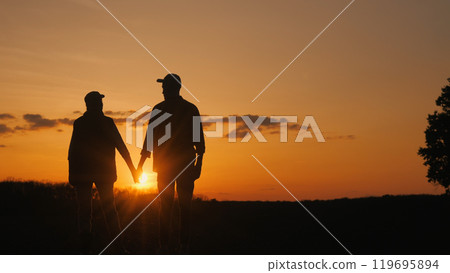 Holding hands, a couple watches the sunset over the grassy field. Holding hands, a couple watches the sunset over the grassy field. 119695894