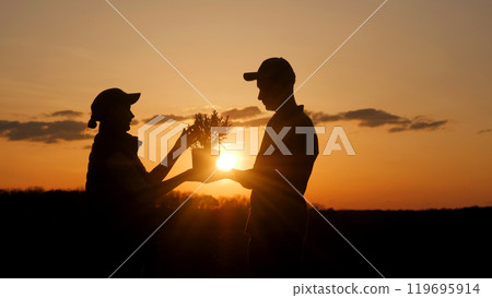 At sunset, a couple of farmers stand in a field, each holding a seedling in their hands. At sunset, a couple of farmers stand in a field, each holding a seedling in their hands. 119695914