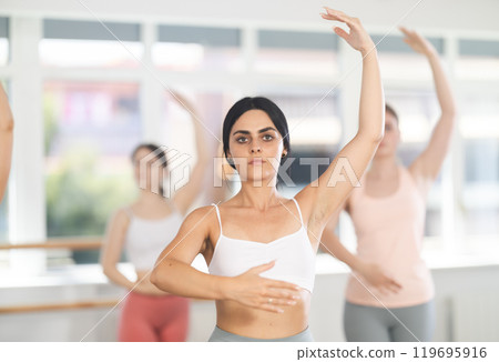 Portrait of pretty young girl dancer exercising various ballet moves during training with group of sporty people in training room 119695916