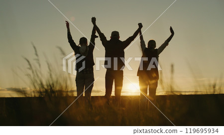 Against the backdrop of the setting sun, a family of farmers raises their hands in a confident sign of success while standing in their field. 119695934