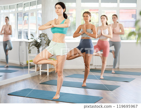Young girl teaches yoga lesson, shows students technique of performing pose of tree asana Young girl teaches yoga lesson, shows students technique of performing pose of tree asana 119695967