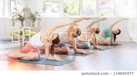 Women exercising Hatha yoga in fitness studio, sitting on mat and doing stretching in Parivrtta Janu Sirsasana Women exercising Hatha yoga in fitness studio, sitting on mat and doing stretching in Parivrtta Janu Sirsasana 119695977