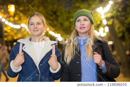 Positive woman walking with teenage daughter along evening city street 119696035
