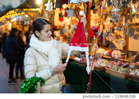 Joyful young woman chooses Santa Claus hat at street market Joyful young woman chooses Santa Claus hat at street market 119696106