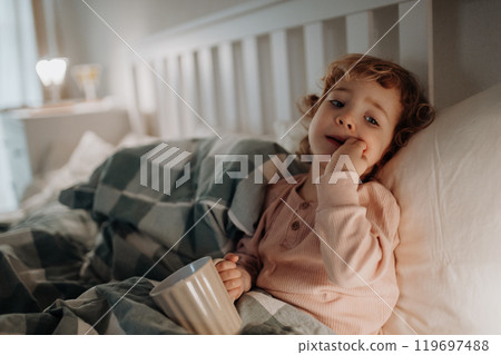 Little girl lying in the bed, holding tea cup. Sick girl with cold is drinking herbal tea. 119697488